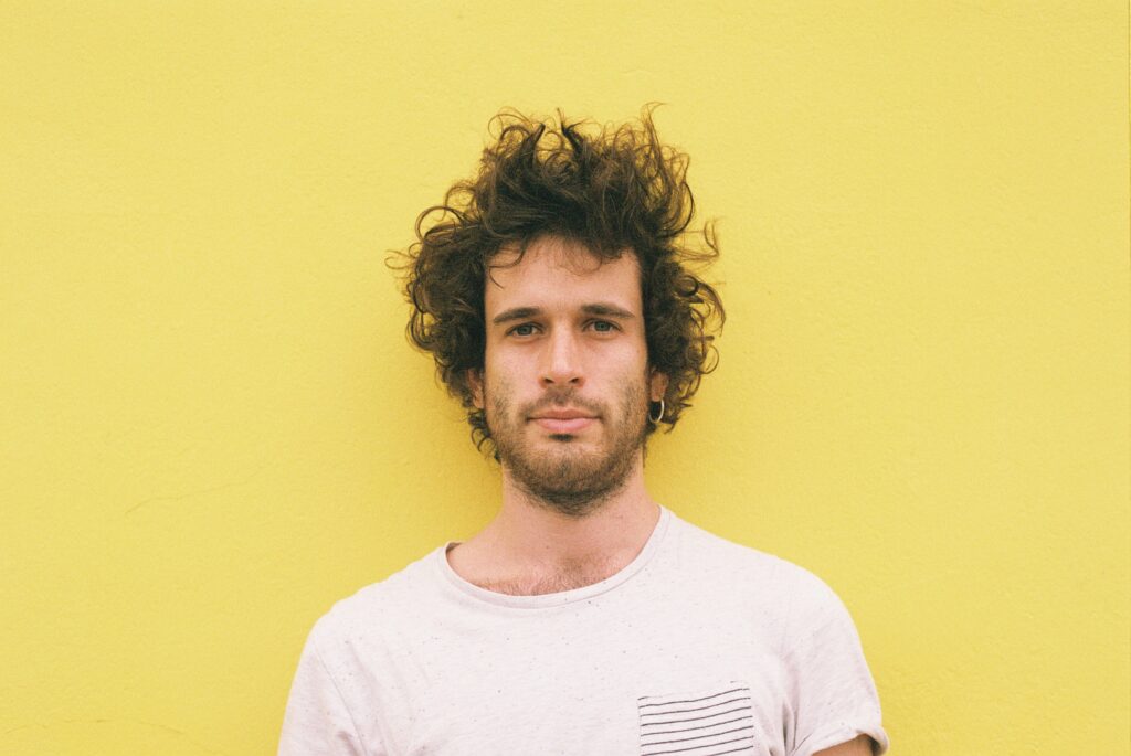Portrait d’un artisan ébéniste en t-shirt clair, devant un mur jaune uni, regardant l’objectif avec une expression calme.